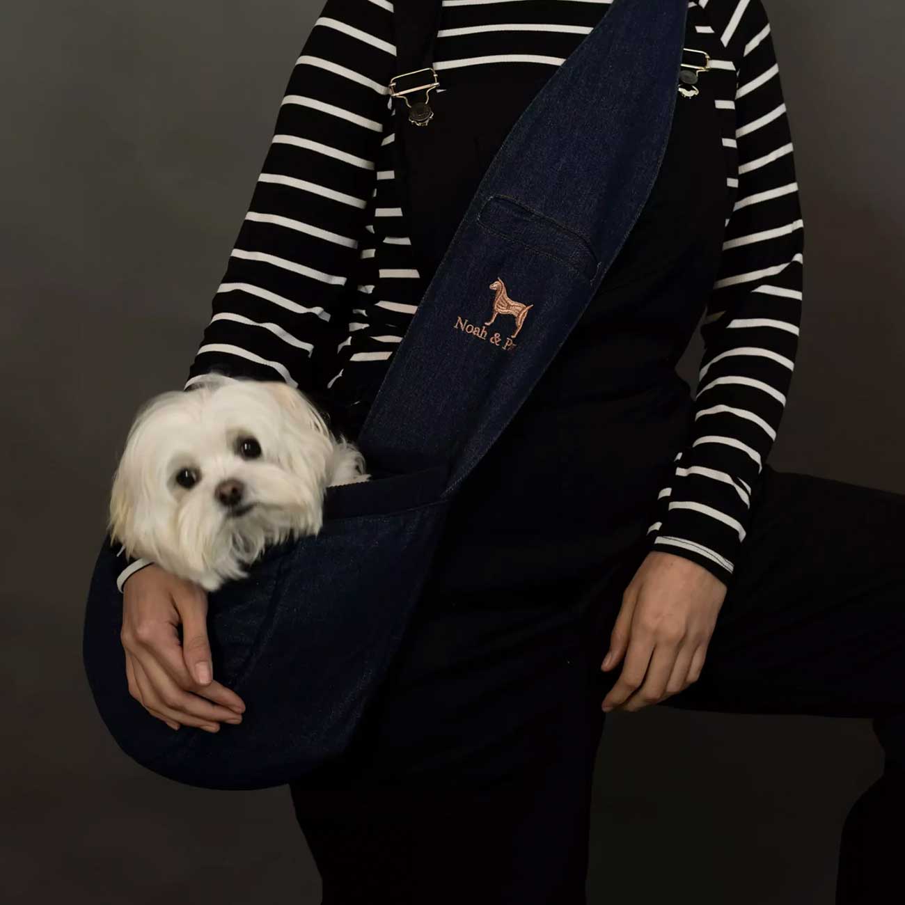 Person holding a small white dog with a dark blue denim pet carrier featuring a logo on a dark background
