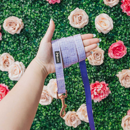 Hand holding a purple dog leash with a brand logo against a floral background