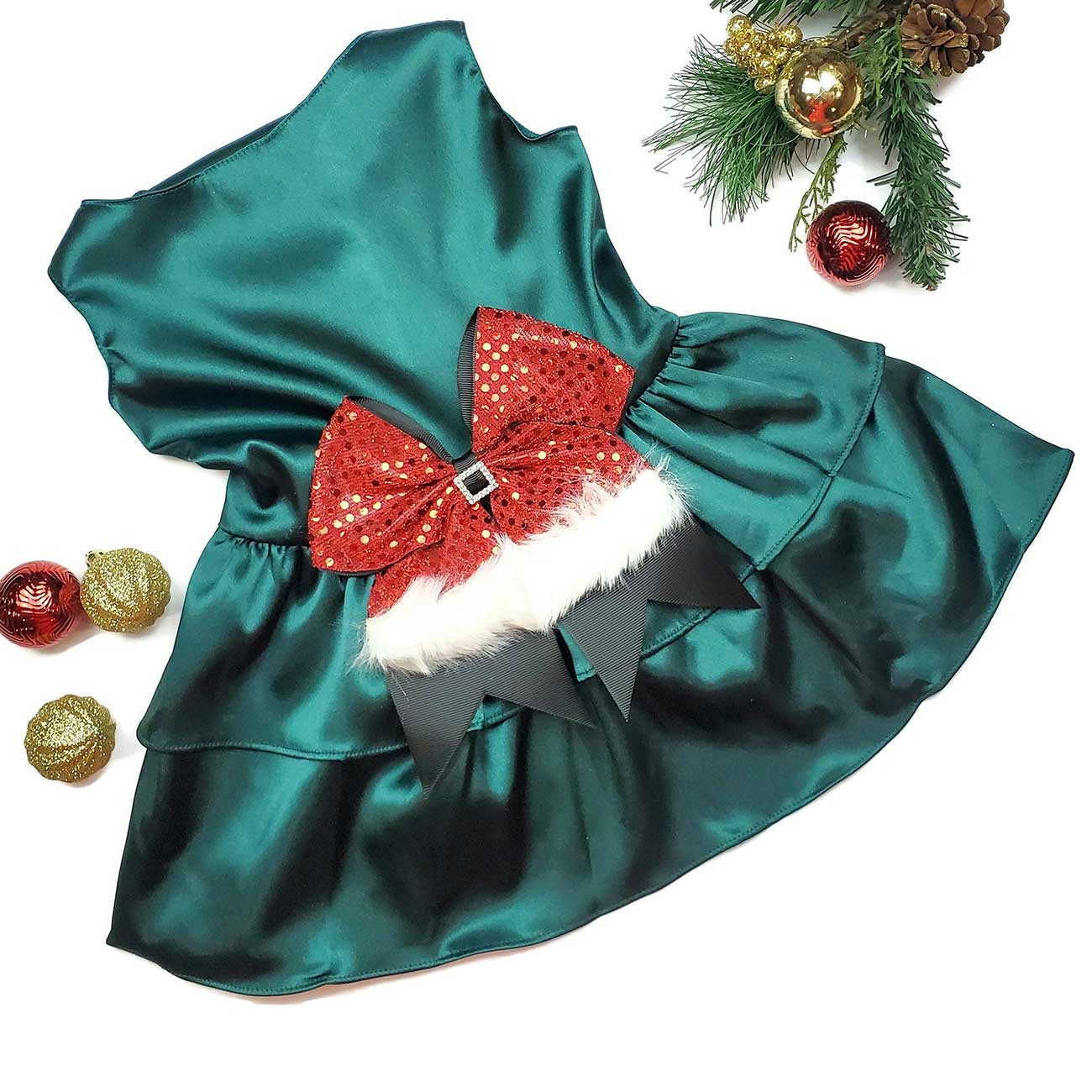 Green satin dog dress with a red sequin bow on a white background with Christmas decorations.