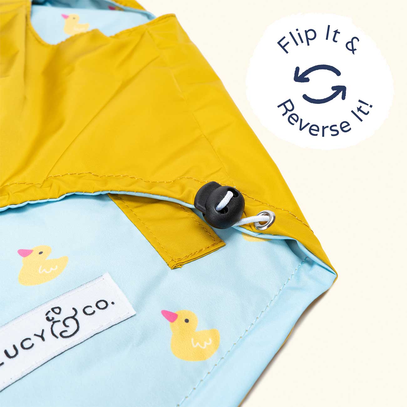 Close-up of a reversible dog's raincoat with yellow and blue sections, featuring a rubber duck design and the brand 'Lucy & Co.'.