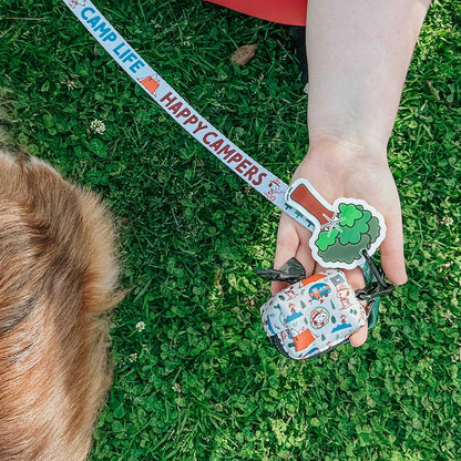 Person holding a dog leash with 'Happy Campers' and a tree-shaped dog waste bag holder on grass