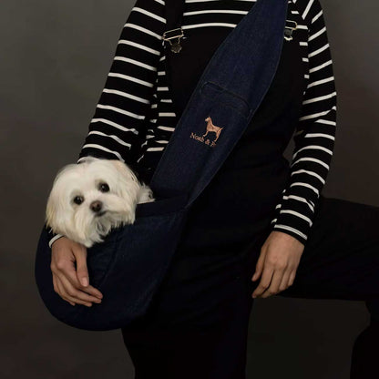 Person holding a small white dog with a dark blue denim pet carrier featuring a logo on a dark background