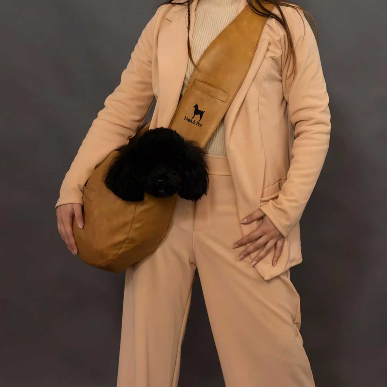 Person wearing a beige outfit with a tan dog bag carrier holding a black dog, against a dark background