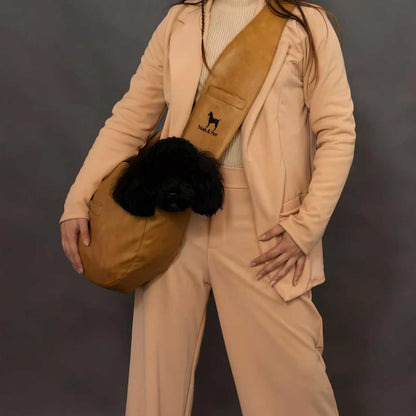 Person wearing a beige outfit with a tan dog bag carrier holding a black dog, against a dark background